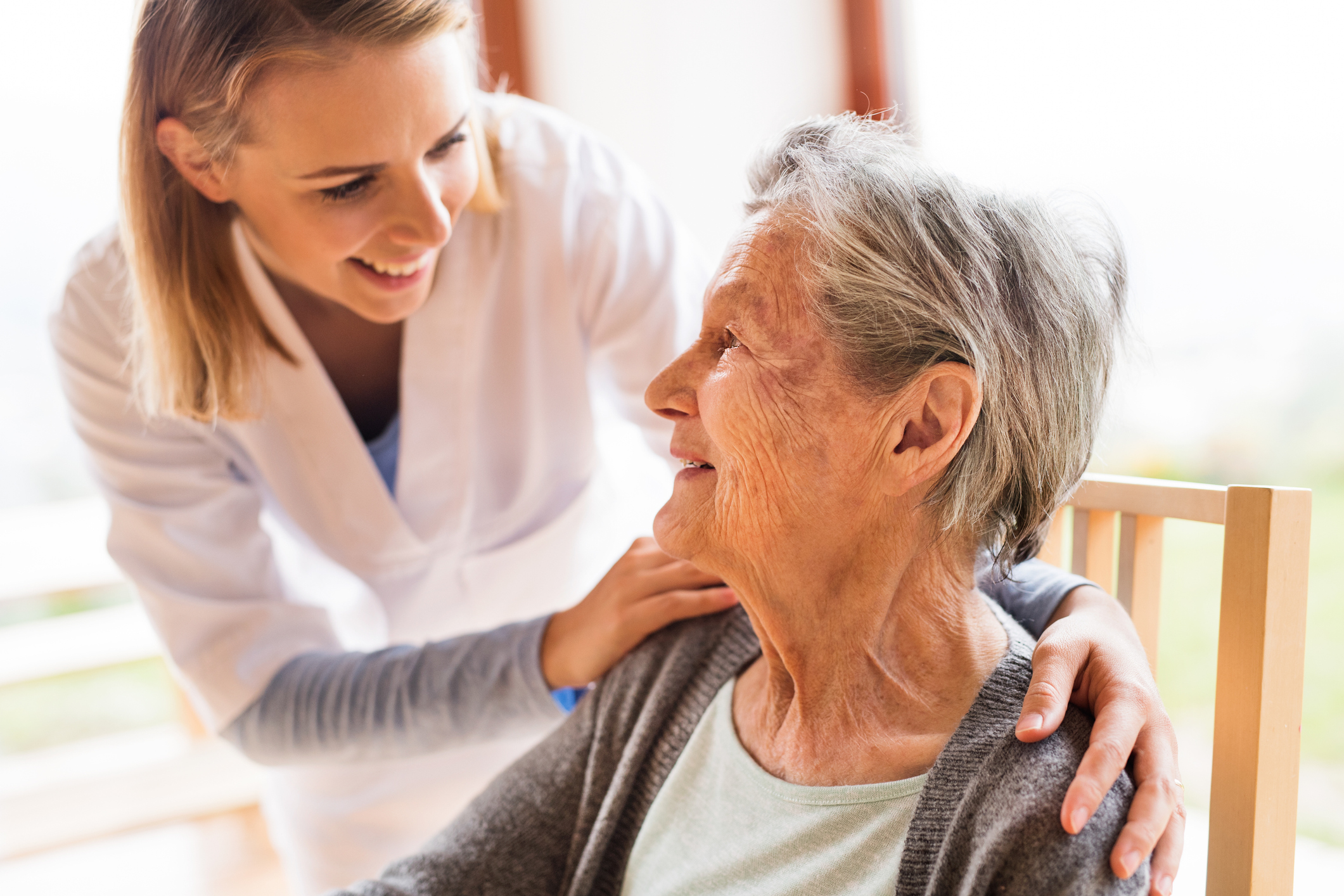 A young female support worker smiles at and touches the shoulder of an elderly woman in a wheelchair.