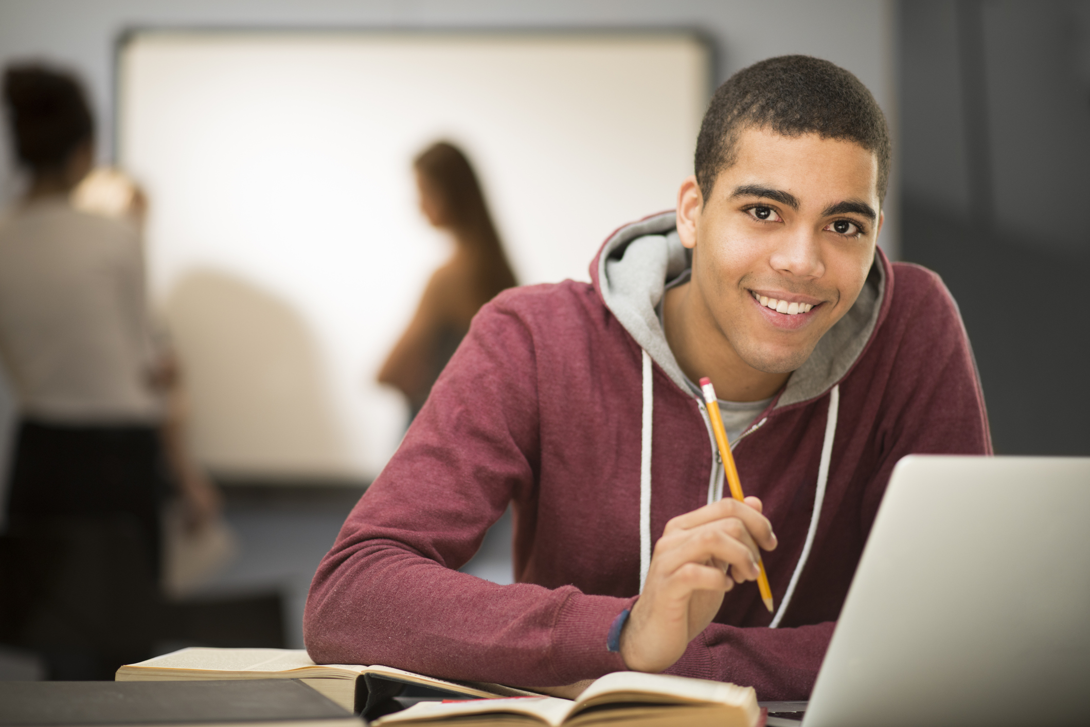 Young man wearing a red hoodie looks up from his laptop, smiles to camera. 