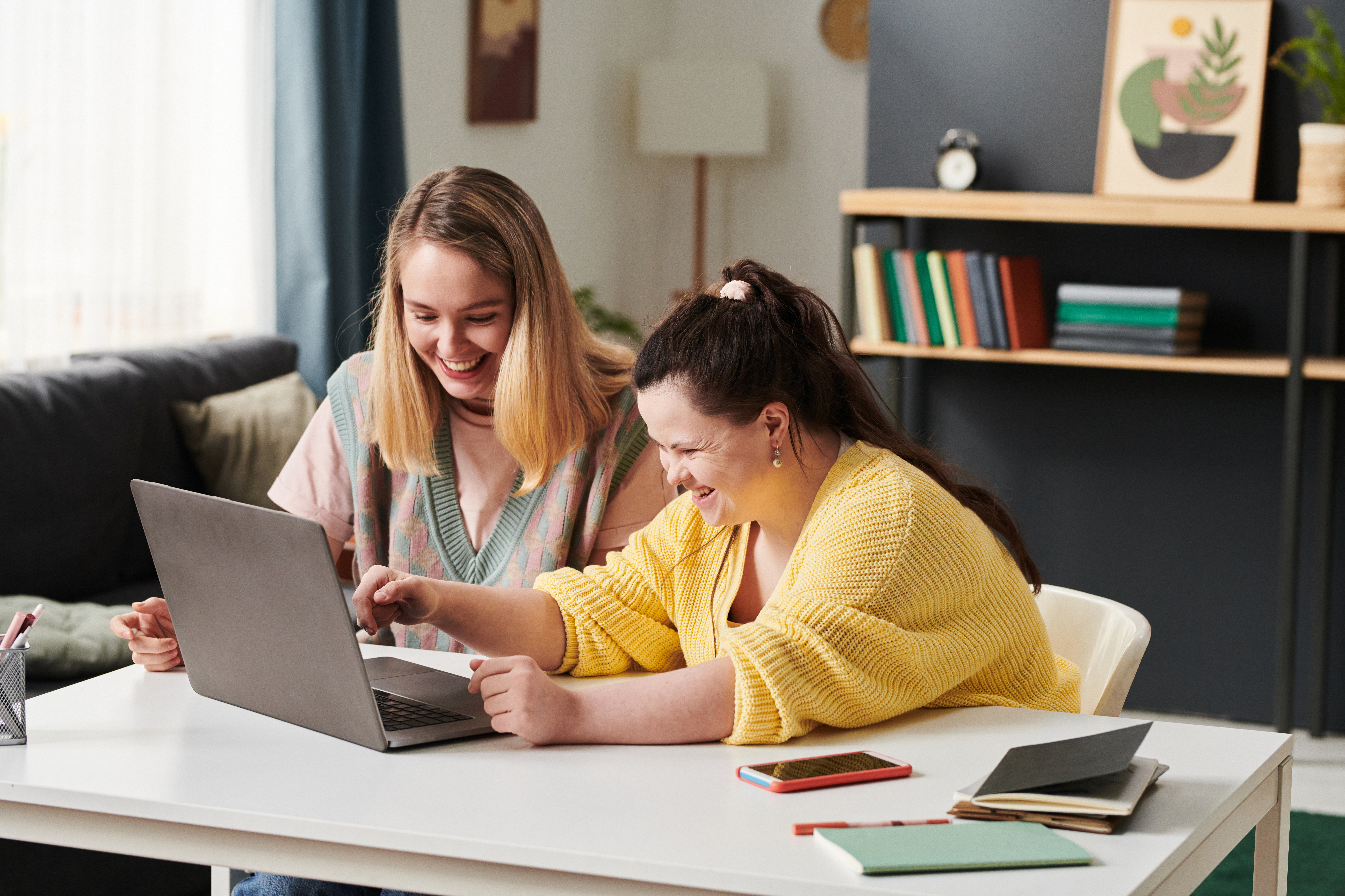 Two women smiling and looking at a laptop together at a table, one helping the other.